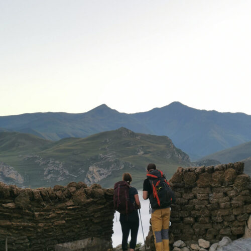 Two hikers passing a stone wall on a ridge path with hazy mountains behind, Guba-Gusar, Azerbaijan