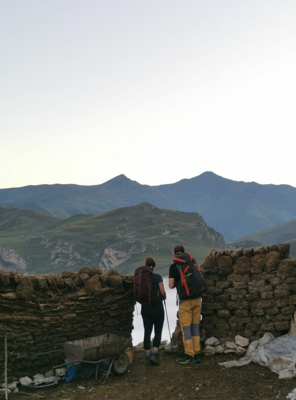 Two hikers passing a stone wall on a ridge path with hazy mountains behind, Guba-Gusar, Azerbaijan