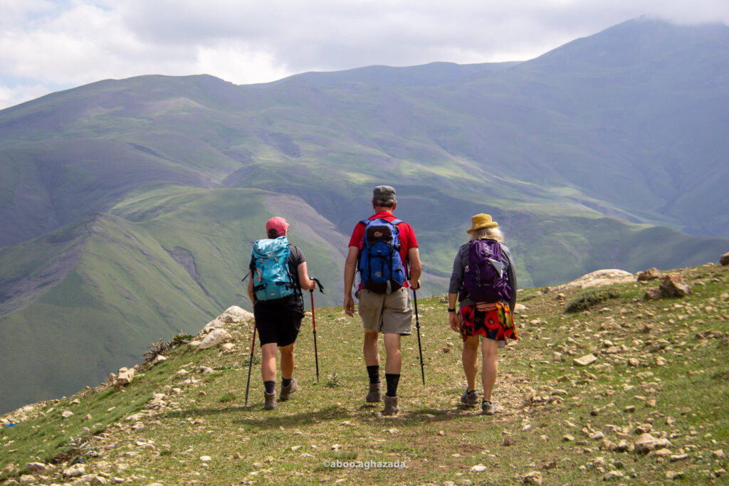 Three hikers with backpacks walking a grassy ridge trail above green valleys, Azerbaijan
