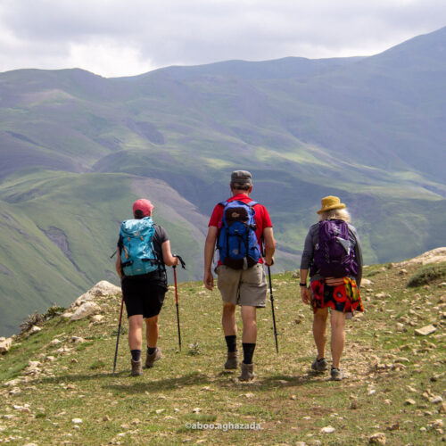 Three hikers with backpacks walking a grassy ridge trail above green valleys, Azerbaijan