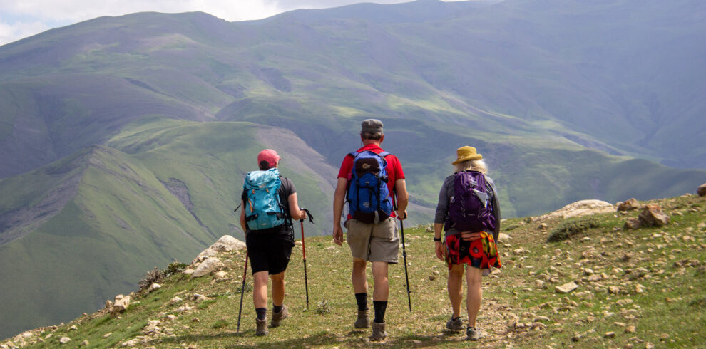 Three hikers with backpacks walking a grassy ridge trail above green valleys, Azerbaijan